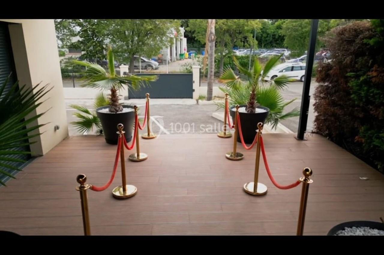 Entrée extérieure avec sol en bois, plantes en pot et barrières dorées reliées par des cordons rouges.