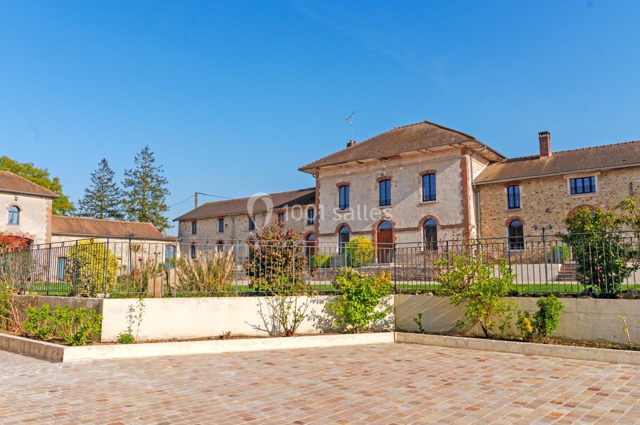 Bâtiment ancien en pierre avec toit en tuiles, entouré d'un jardin et d'une cour pavée sous un ciel bleu.
