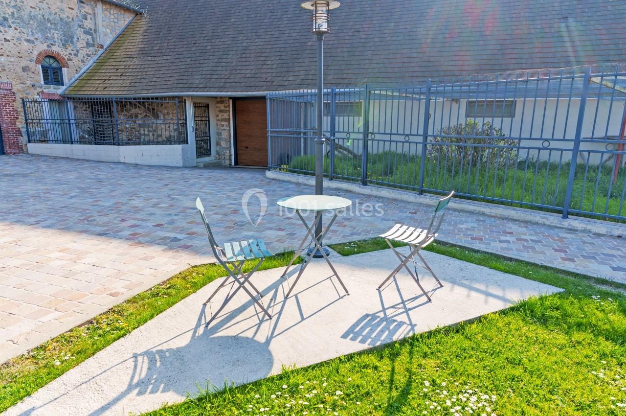 Deux chaises et une table en métal sur une terrasse pavée, près d'une pelouse et d'un bâtiment en pierre.