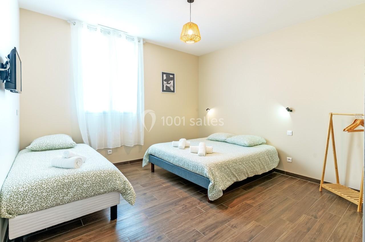 Chambre lumineuse avec un lit double, un lit simple, des draps verts, un portant et un sol en parquet.