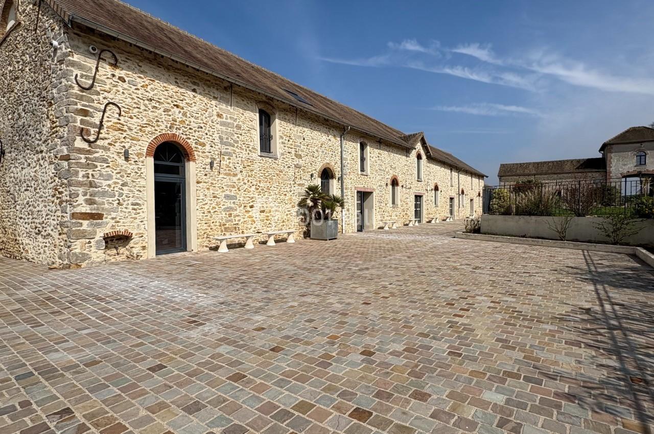 Bâtiment en pierre avec toit en tuiles, grande cour pavée et ciel dégagé en arrière-plan.