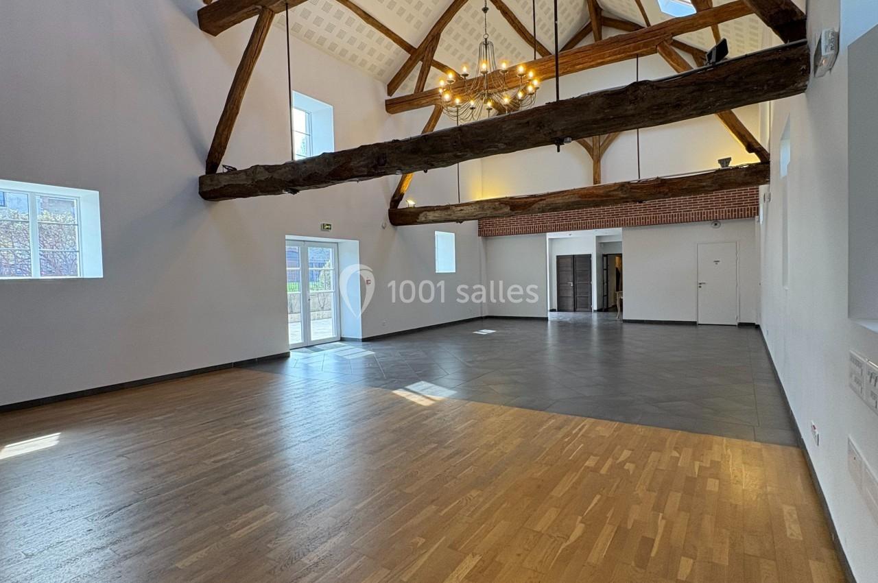Salle spacieuse avec poutres apparentes, parquet en bois clair, murs blancs et grandes fenêtres laissant entrer la lumière…