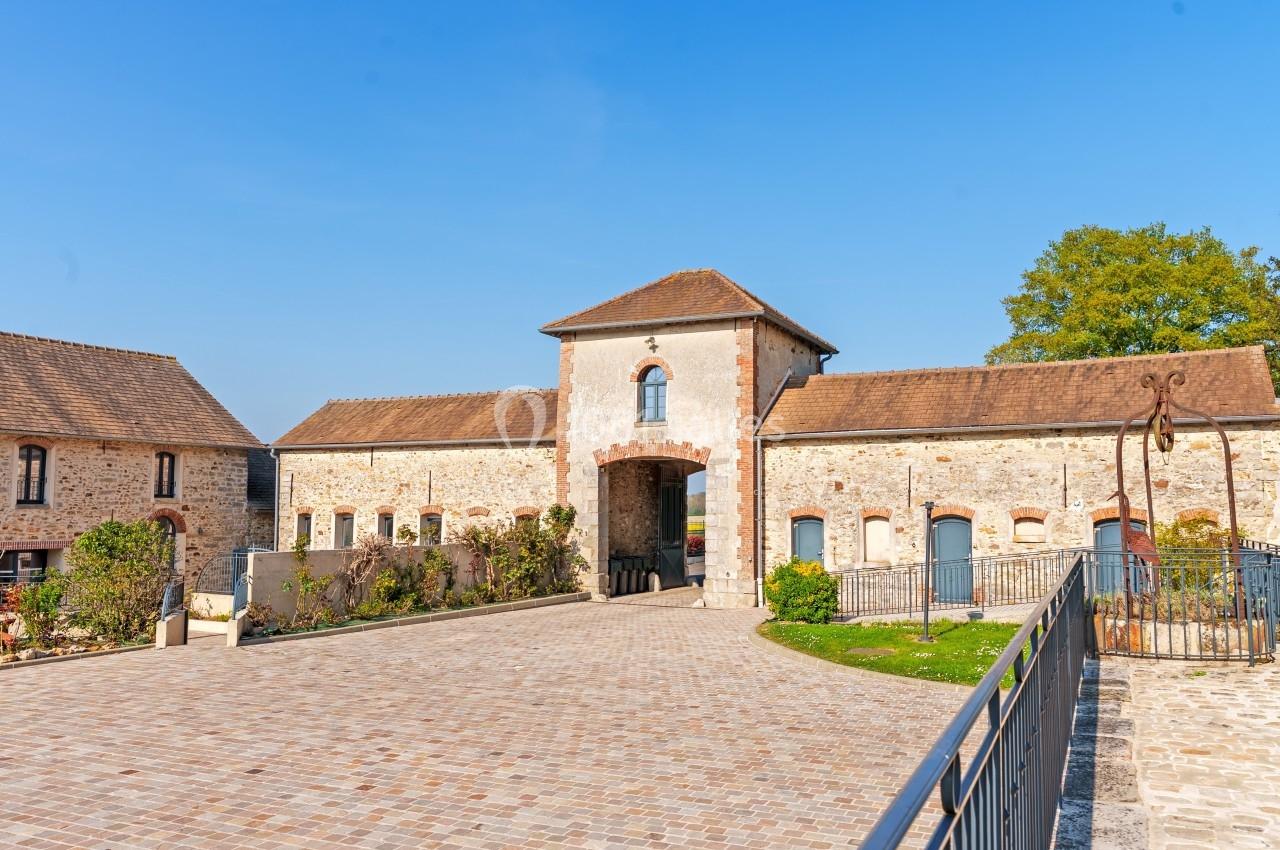 Cour pavée avec un bâtiment en pierre et une grande porte centrale sous un toit en tuiles, entouré de verdure.