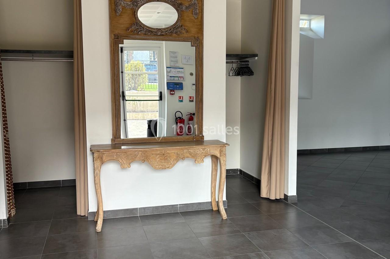 Salle vide avec sol en carrelage gris, miroir encadré au-dessus d'une console en bois sculpté et rideaux beige aux murs.