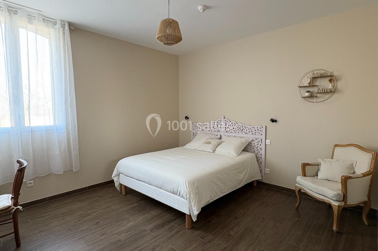 Chambre lumineuse avec lit double, parquet foncé, fauteuils et fenêtre avec rideaux blancs.