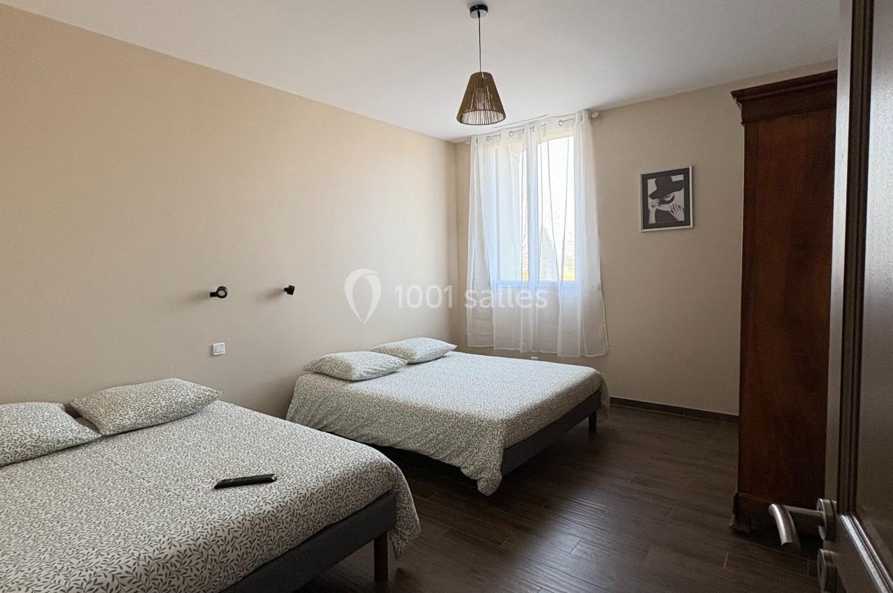 Deux lits doubles avec linge de lit clair dans une chambre lumineuse au sol en bois sombre, avec une armoire et une fenêtre.