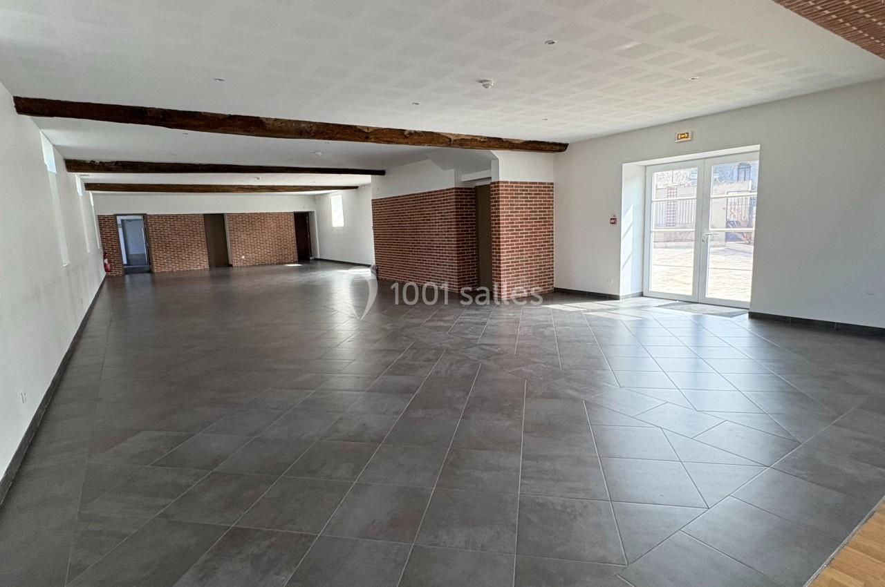 Salle spacieuse avec sol en carrelage gris, murs blancs, poutres apparentes et briques, éclairée par des fenêtres.