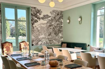 Salle de restaurant lumineuse avec tables dressées, banquettes vertes, chaises blanches et vue sur un jardin arboré.