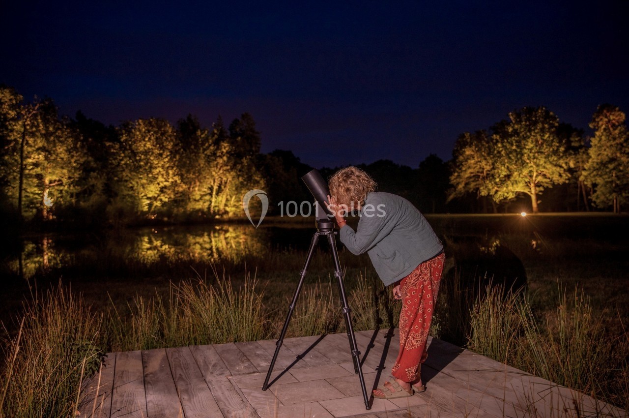 Une personne observe le ciel nocturne à l'aide d'un télescope sur une plateforme en bois près d'un étang éclairé.
