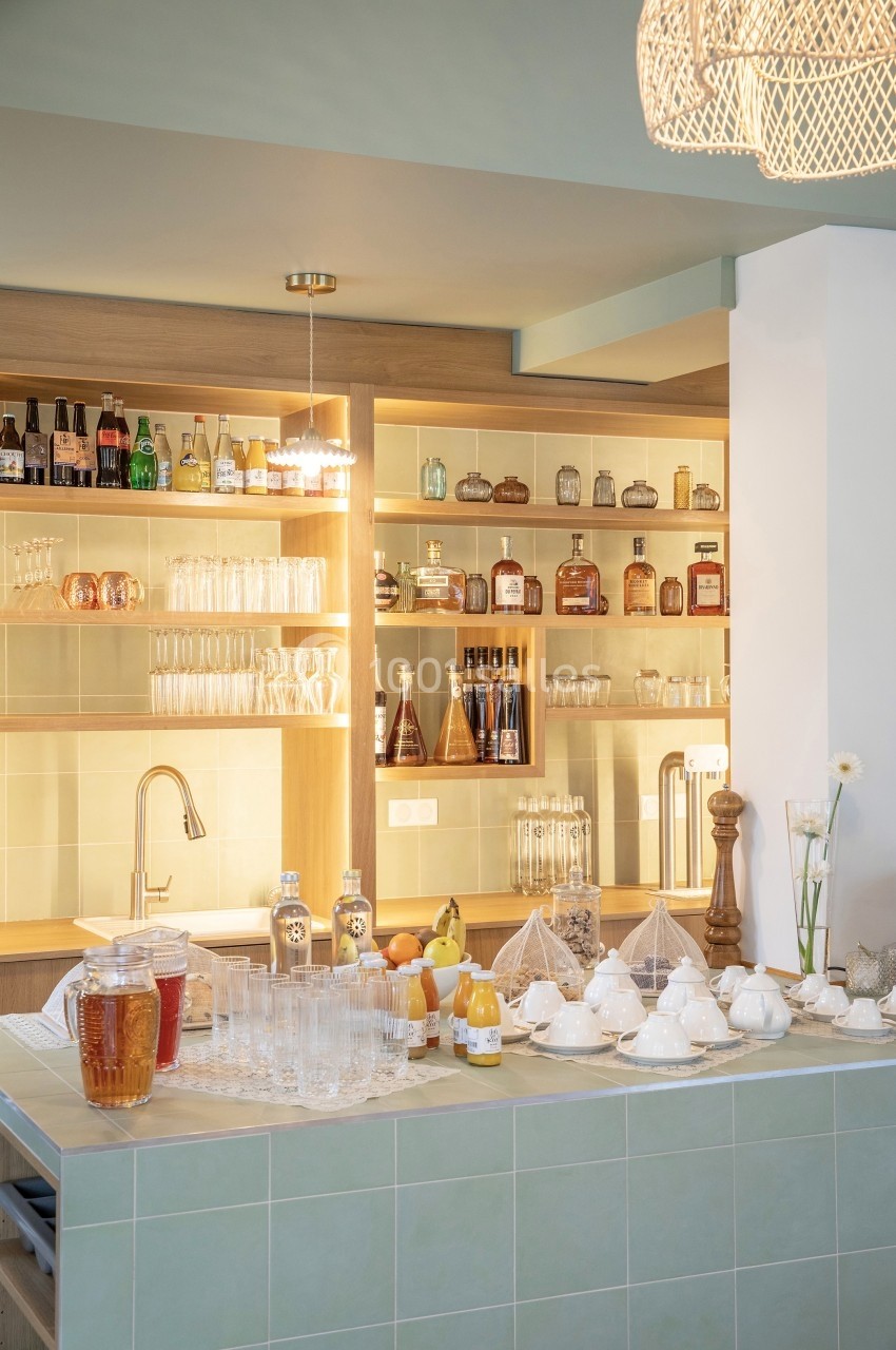 Un comptoir de cuisine avec boissons, verres, théières et étagères remplies de bouteilles en arrière-plan.