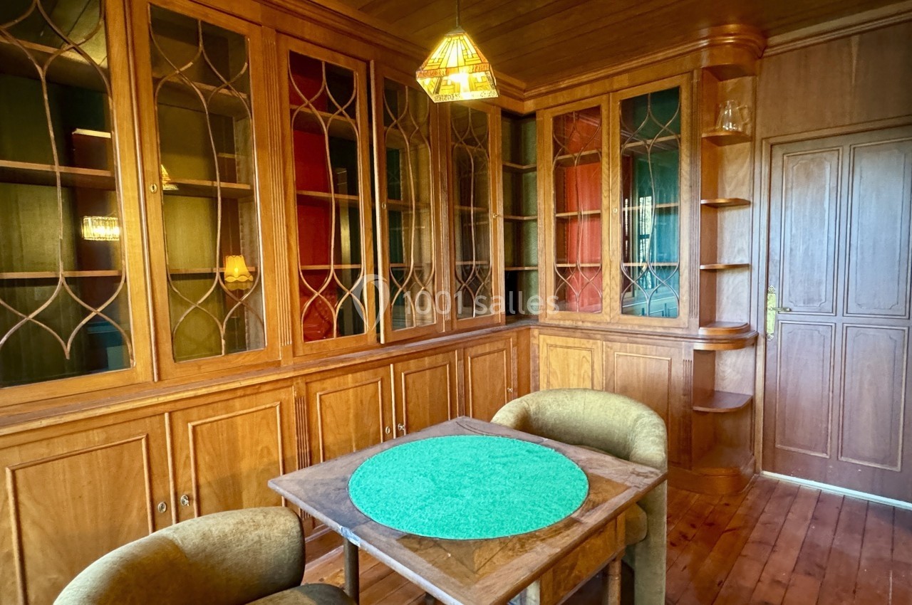 Salle à manger avec boiseries, étagères vitrées, table carrée avec napperon vert et deux fauteuils rembourrés.