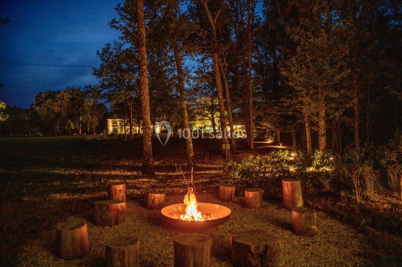 Feu de camp entouré de rondins de bois dans une clairière boisée, avec des lumières de maison en arrière-plan.