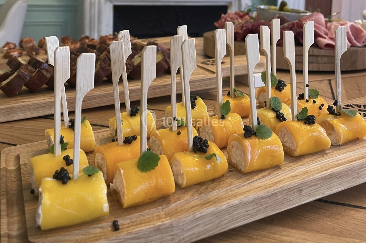 Plateau de bouchées roulées au fromage et mangue, décorées de caviar et de feuilles, sur une table en bois.