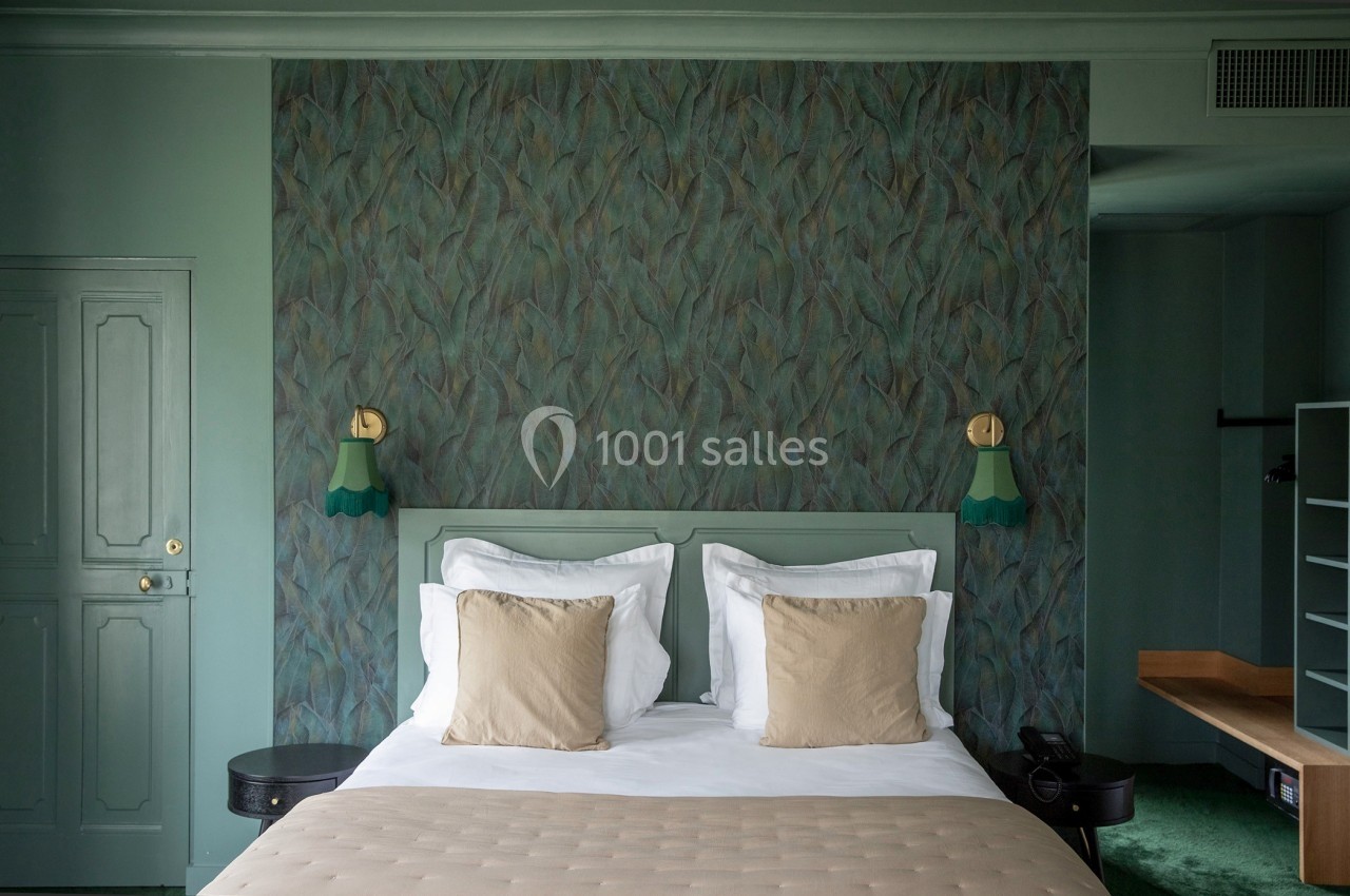 Lit double avec coussins beiges, encadré par des lampes murales vertes, dans une chambre aux tons verts et mur texturé.