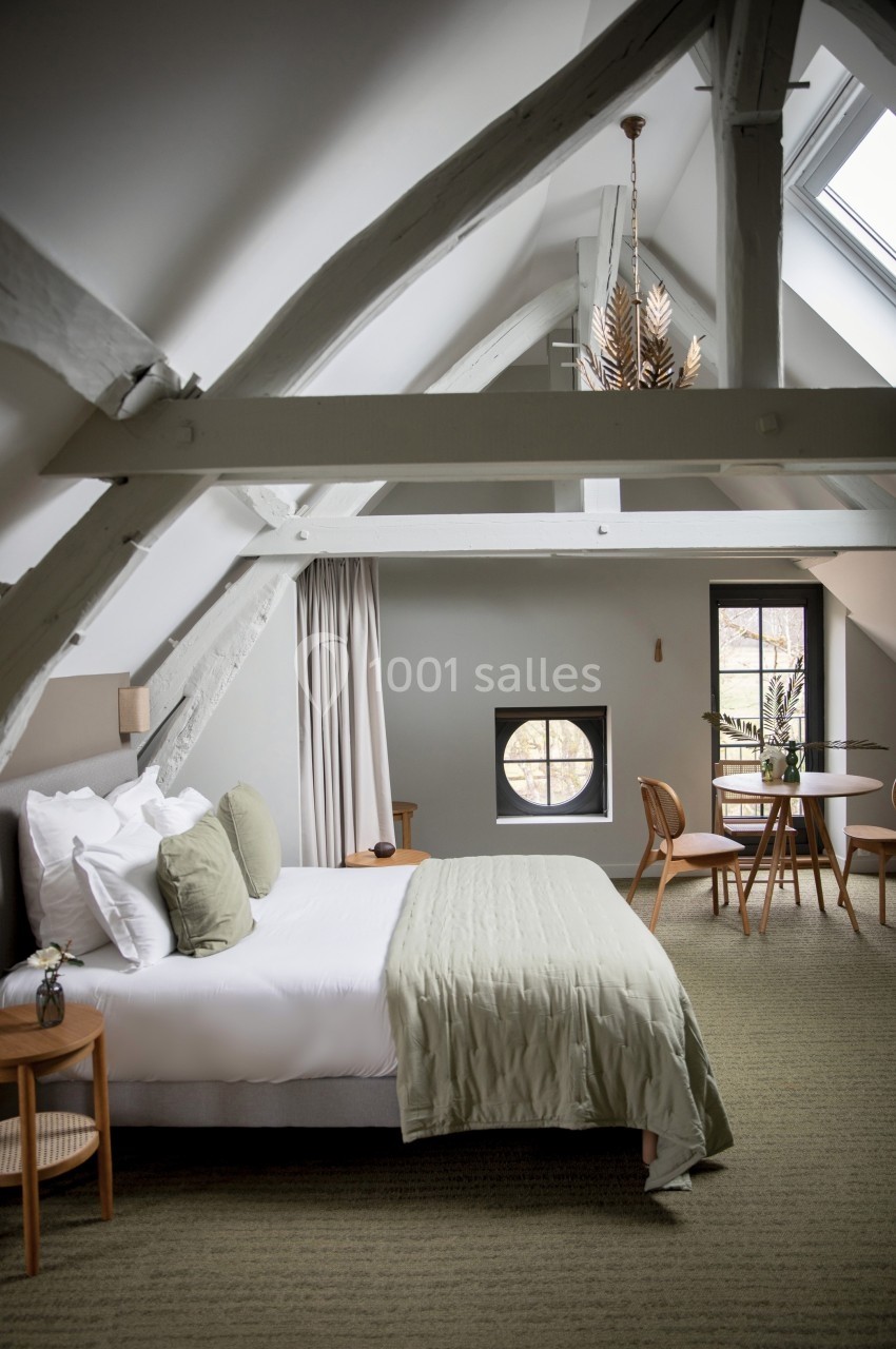 Chambre lumineuse sous combles avec poutres apparentes, lit double, table, chaises et fenêtre ronde.