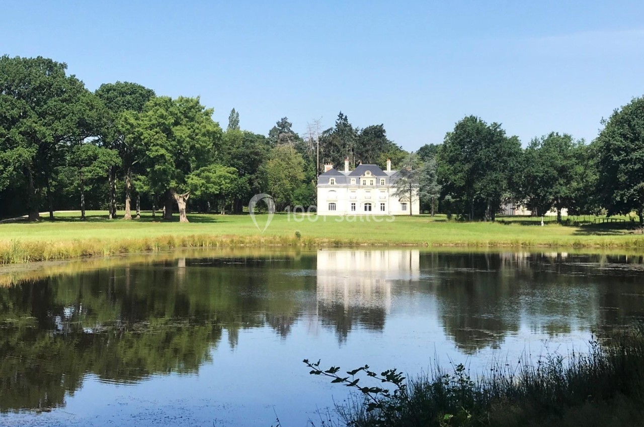 Vue d'un château blanc entouré d'arbres, reflété dans un étang sous un ciel bleu.