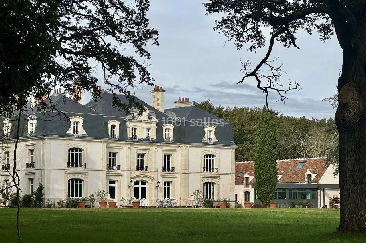Façade d'un manoir blanc entouré d'arbres, avec une pelouse et une dépendance en arrière-plan.