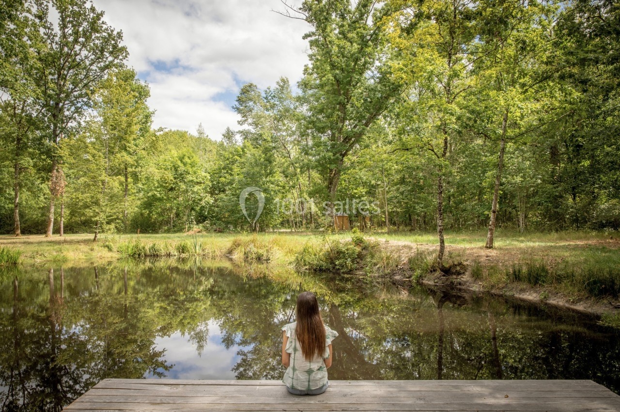 Une femme assise sur un ponton en bois contemple un étang entouré d'arbres sous un ciel partiellement nuageux.