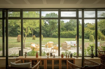 Salle de restaurant lumineuse avec tables dressées, banquettes vertes, chaises blanches et vue sur un jardin arboré.