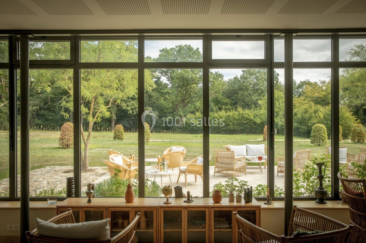 Vue depuis une pièce lumineuse sur une terrasse aménagée avec des fauteuils en rotin et un jardin verdoyant en arrière-plan.