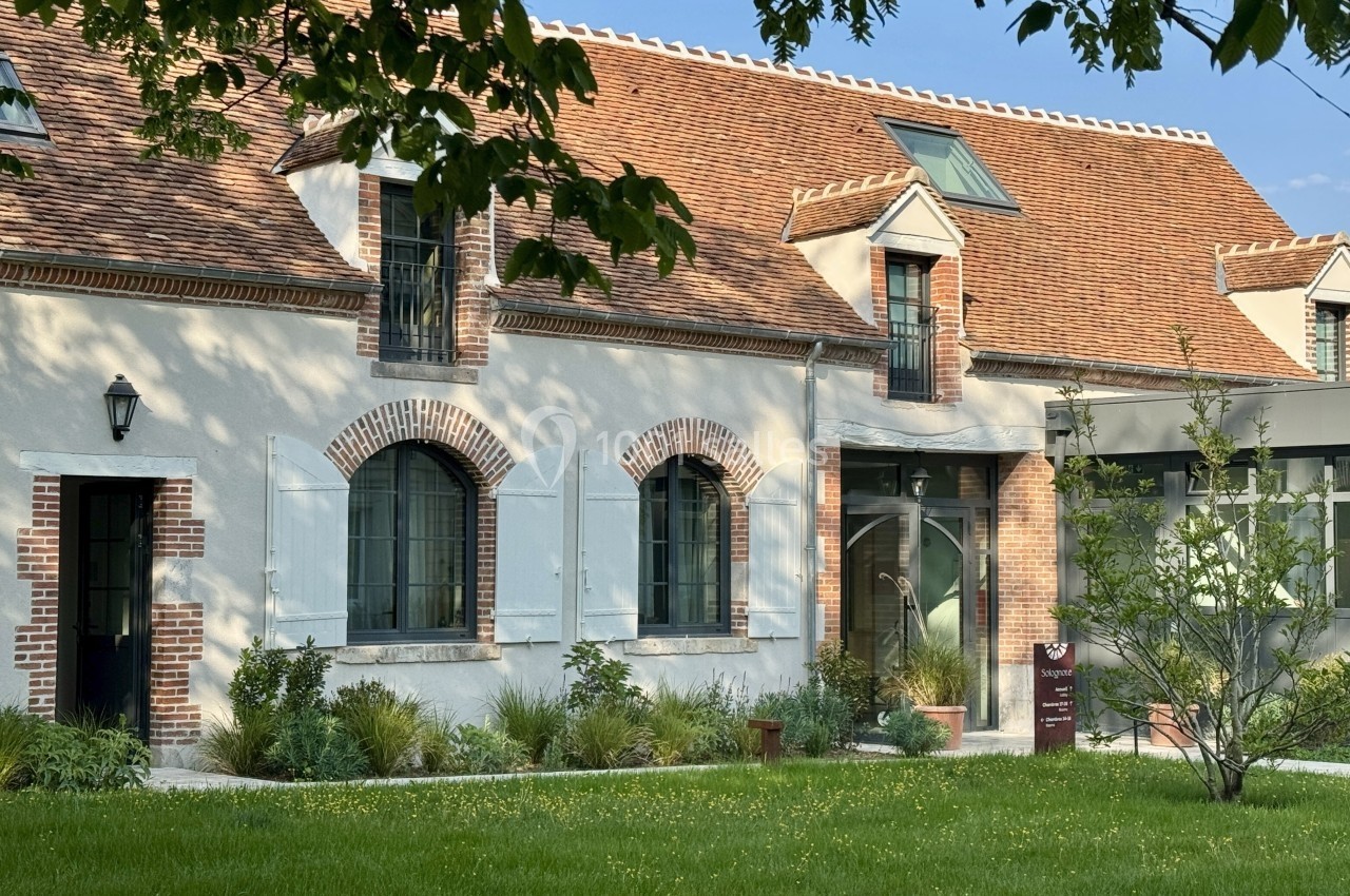 Façade d'une maison en pierre avec toit en tuiles, volets blancs, jardin verdoyant et arbres au premier plan.