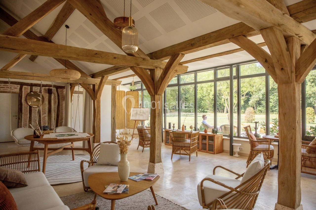 Salle lumineuse avec poutres en bois, mobilier en rotin et grandes baies vitrées donnant sur un jardin verdoyant.