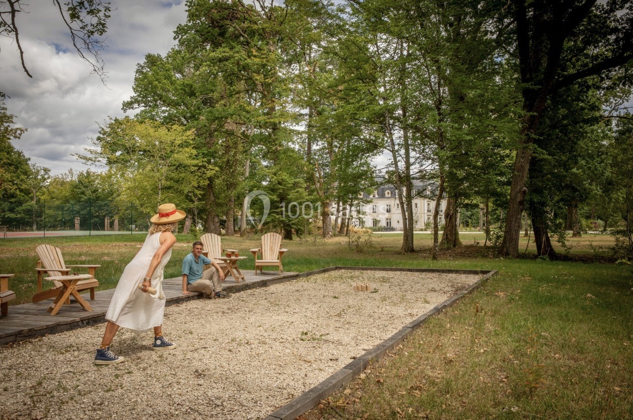 Deux personnes jouent à la pétanque sur un terrain en gravier, entouré d'arbres et avec un bâtiment en arrière-plan.
