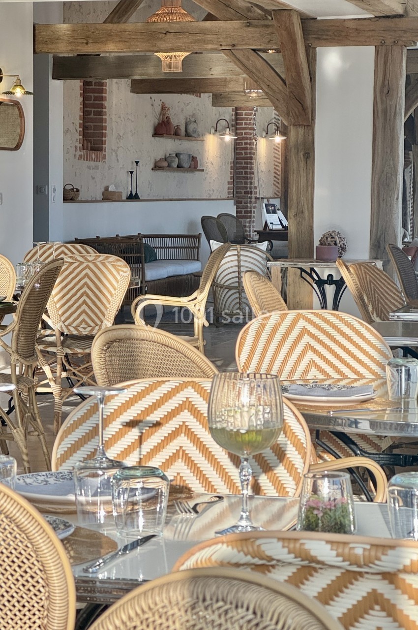 Salle de restaurant lumineuse avec chaises en rotin à motifs, tables dressées et poutres apparentes en bois.