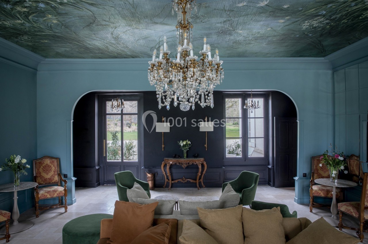 Salon élégant avec un plafond peint, un lustre en cristal, des fauteuils verts et des fenêtres donnant sur un jardin.