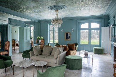 Salle de restaurant lumineuse avec tables dressées, banquettes vertes, chaises blanches et vue sur un jardin arboré.