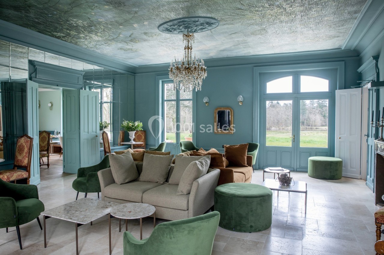 Salon élégant avec murs bleus, mobilier vert et beige, lustre en cristal, grandes fenêtres donnant sur un jardin.