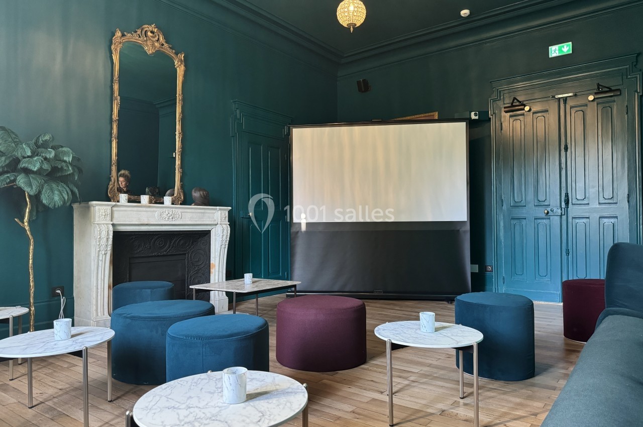 Salle élégante avec murs sombres, cheminée blanche, tables en marbre, poufs colorés et écran de projection.