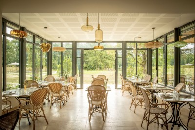 Salle de restaurant lumineuse avec tables dressées, banquettes vertes, chaises blanches et vue sur un jardin arboré.