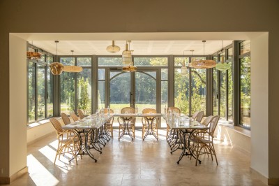 Salle de restaurant lumineuse avec tables dressées, banquettes vertes, chaises blanches et vue sur un jardin arboré.