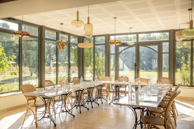 Salle de restaurant lumineuse avec tables dressées, banquettes vertes, chaises blanches et vue sur un jardin arboré.