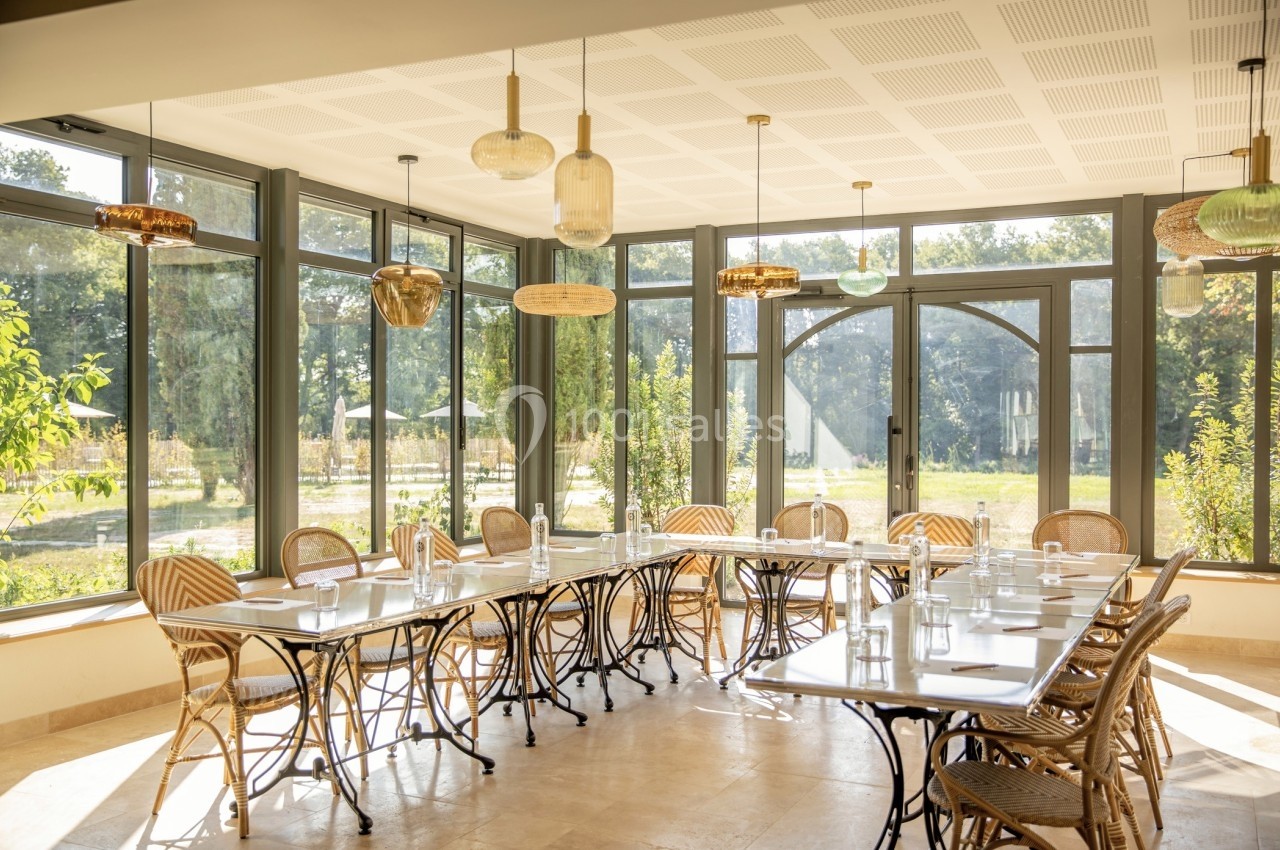 Salle lumineuse avec grandes baies vitrées, tables alignées et chaises en rotin, vue sur un jardin verdoyant.