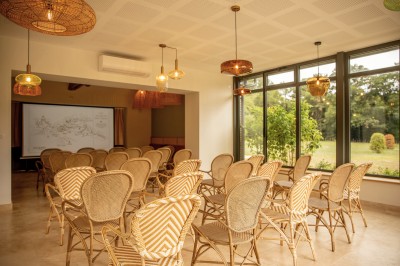 Salle de restaurant lumineuse avec tables dressées, banquettes vertes, chaises blanches et vue sur un jardin arboré.