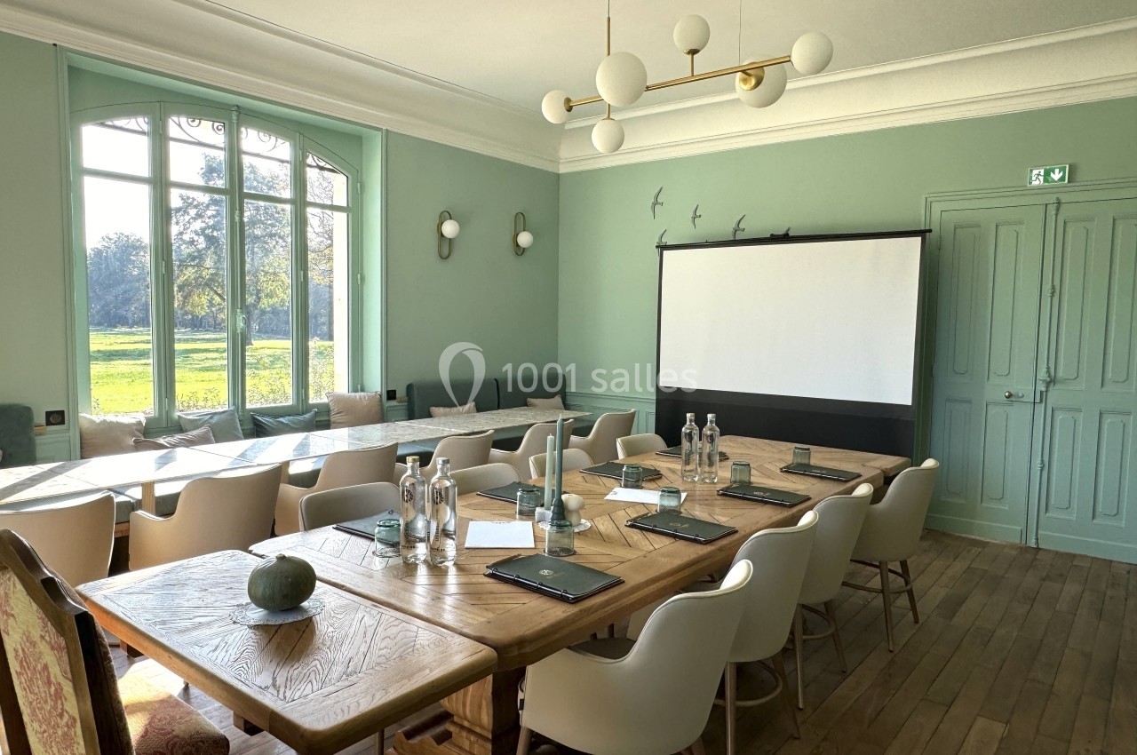 Salle de réunion lumineuse avec grande table en bois, chaises blanches, écran de projection et vue sur un jardin.