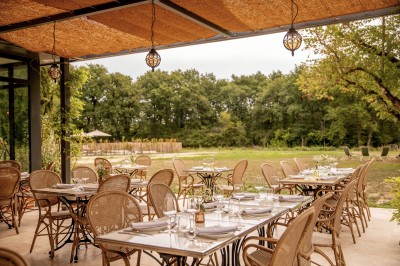 Salle de restaurant lumineuse avec tables dressées, banquettes vertes, chaises blanches et vue sur un jardin arboré.