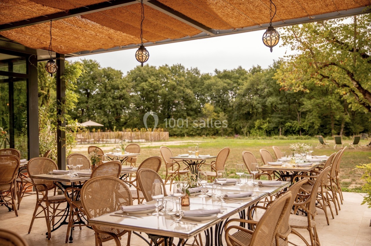 Terrasse d'un restaurant avec des tables dressées, chaises en rotin et vue sur un jardin arboré.