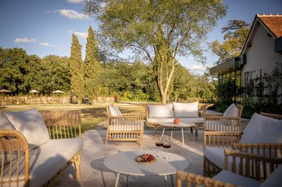 Salle de restaurant lumineuse avec tables dressées, banquettes vertes, chaises blanches et vue sur un jardin arboré.