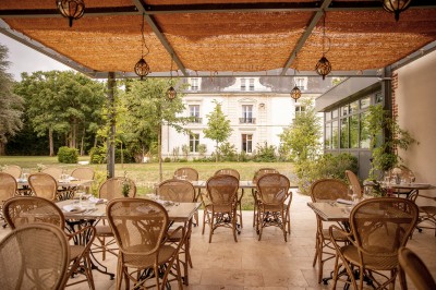 Salle de restaurant lumineuse avec tables dressées, banquettes vertes, chaises blanches et vue sur un jardin arboré.
