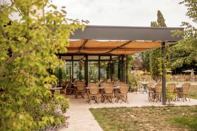 Salle de restaurant lumineuse avec tables dressées, banquettes vertes, chaises blanches et vue sur un jardin arboré.