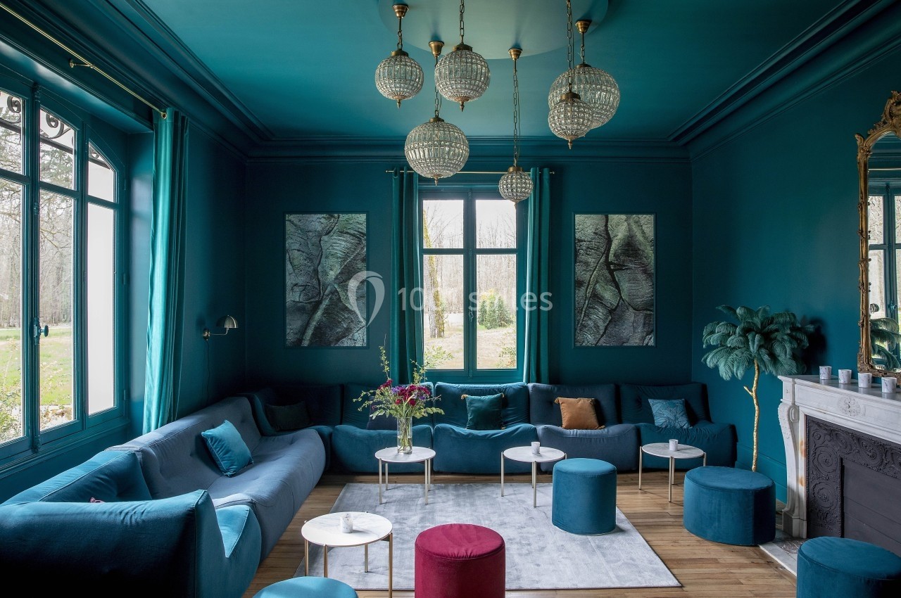 Salon élégant avec murs bleu profond, canapés assortis, tables basses, lustres en cristal et grande fenêtre donnant sur un…