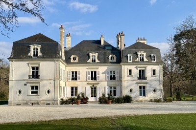 Salle de restaurant lumineuse avec tables dressées, banquettes vertes, chaises blanches et vue sur un jardin arboré.