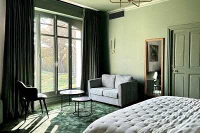 Salle de restaurant lumineuse avec tables dressées, banquettes vertes, chaises blanches et vue sur un jardin arboré.