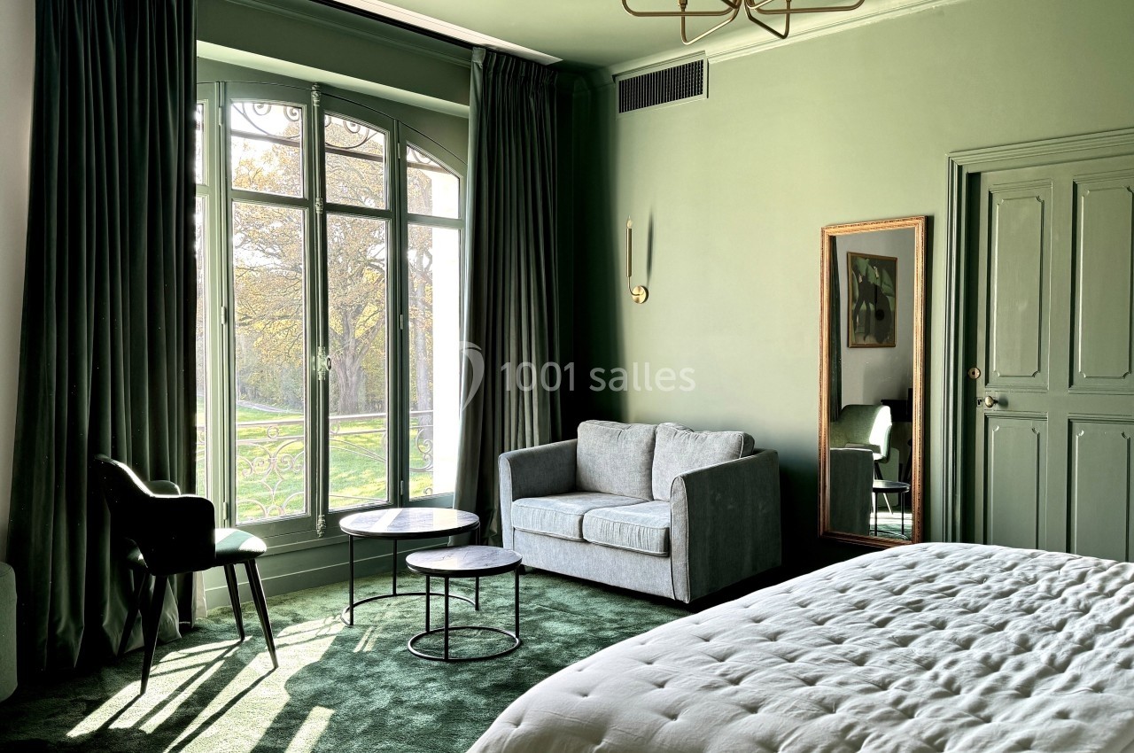 Chambre lumineuse avec lit, canapé, tables basses, grande fenêtre donnant sur un jardin, murs et moquette verts.