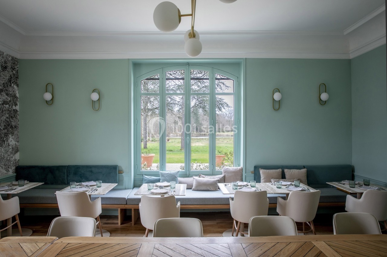 Salle de restaurant lumineuse avec tables dressées, banquettes vertes, chaises blanches et vue sur un jardin arboré.