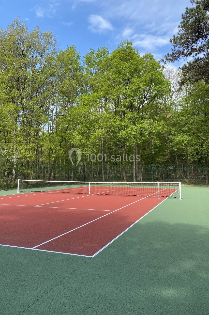 Terrain de tennis extérieur avec revêtement rouge et vert, entouré d'arbres et d'une clôture, sous un ciel partiellement…