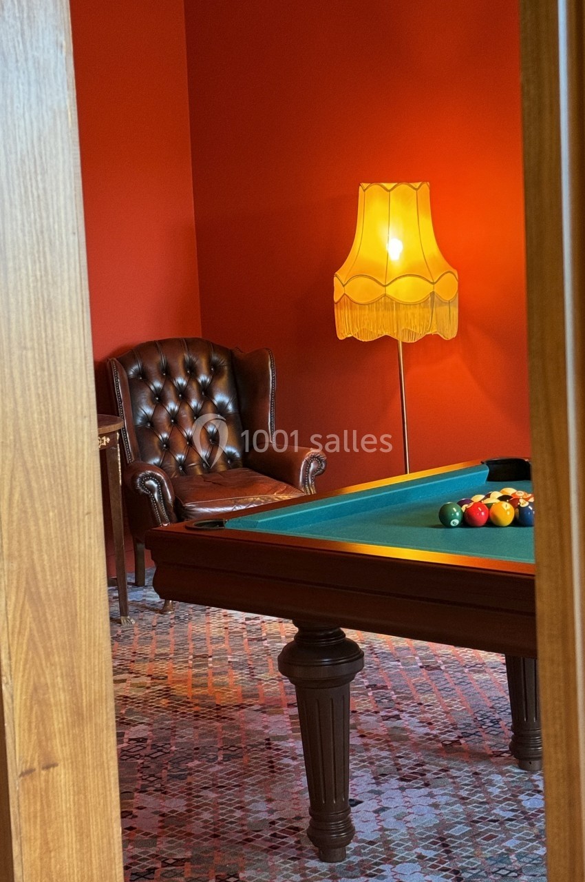 Salle avec un billard, un fauteuil en cuir marron et une lampe rétro éclairant un mur rouge.